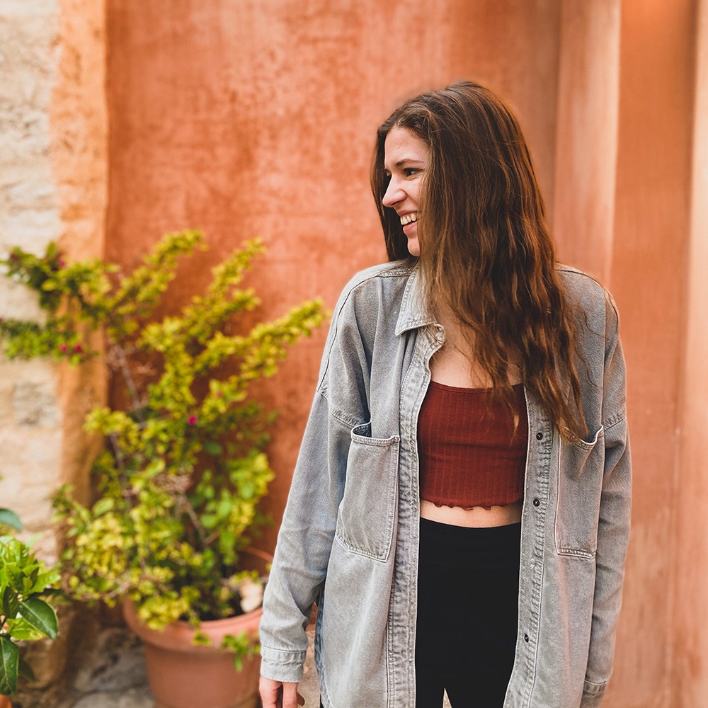 A young woman with long hair stands smiling near a potted plant against a colorful wall.