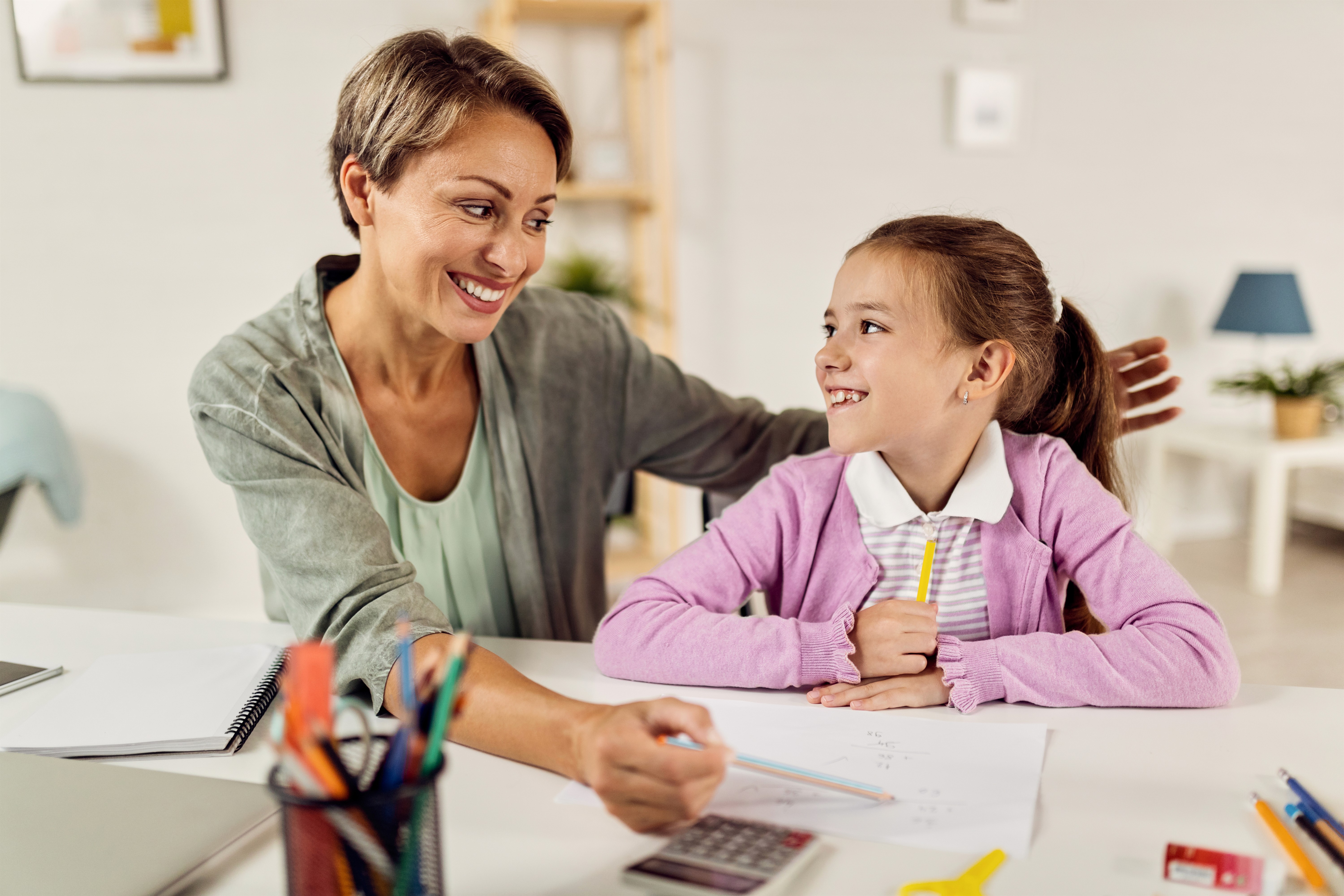 Mulher sorrindo com uma criança, aparentemente estudando educação financeira com uma calculadora e canetas pela mesa