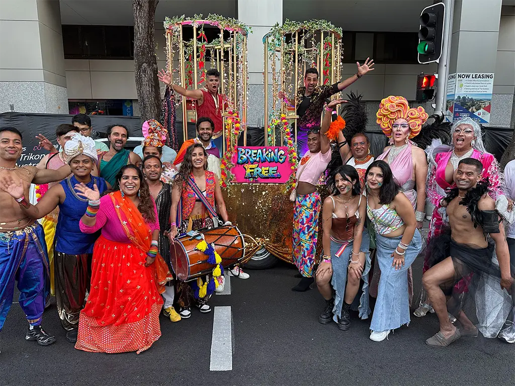 A vibrant group poses in colorful costumes, with smiles and playful gestures. Behind them, a decorated float reads "Breaking Free," conveying joy and celebration.