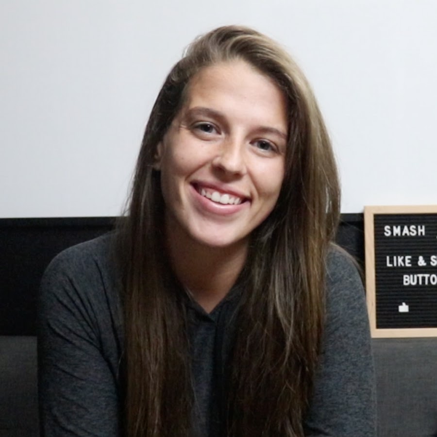 A young woman with long dark hair smiles confidently while crossing her arms in a modern indoor setting.