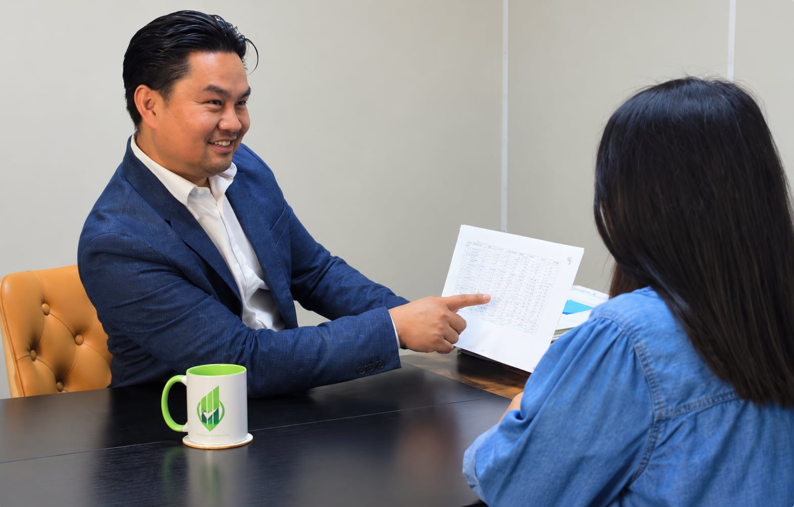 A smiling man in a navy blazer pointing to a printed document while speaking with a woman seated across a desk.