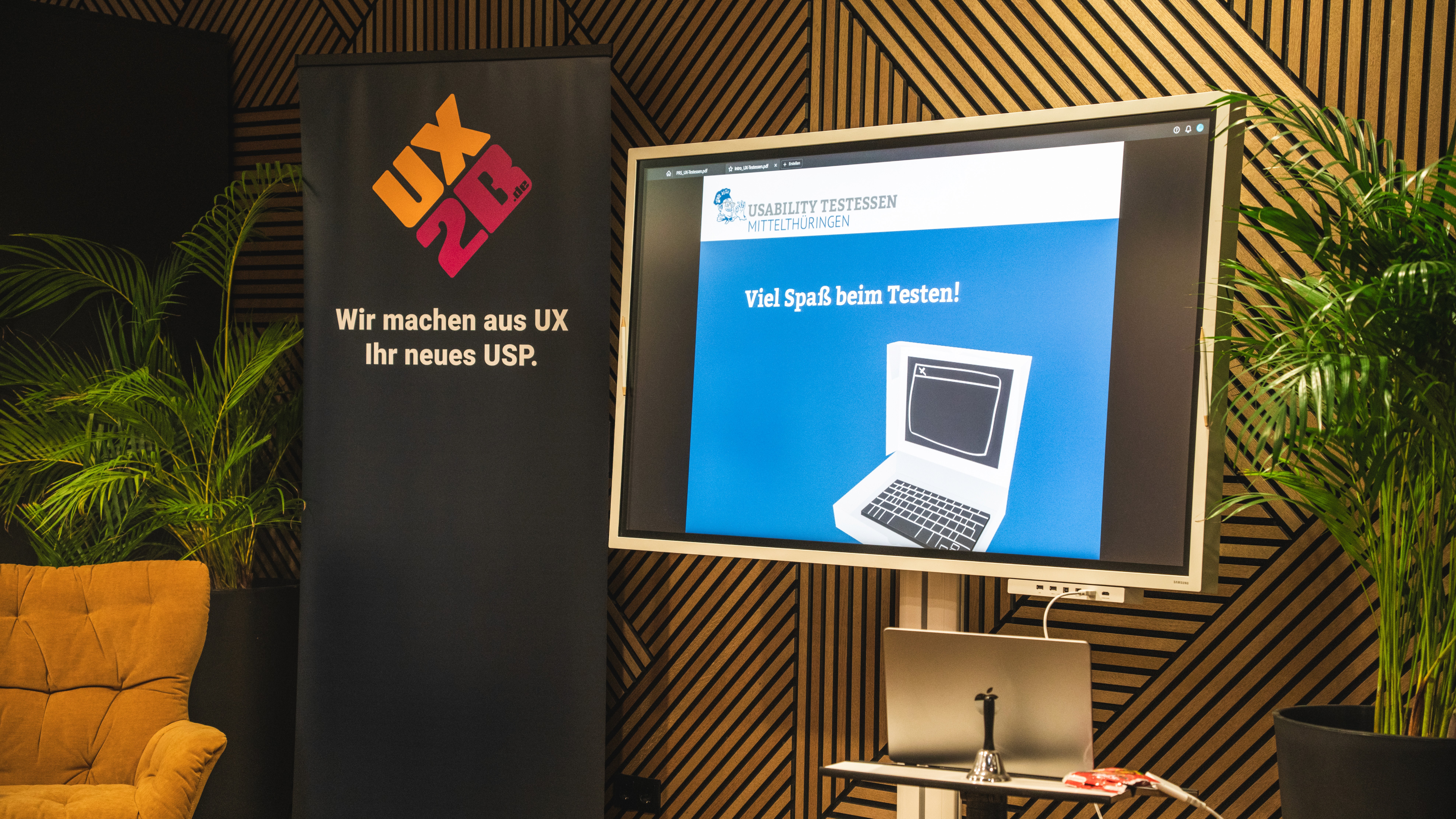 Moderner Raum mit Holzpaneelwand, zwei großen Pflanzen, einem gelben Sessel, einem Banner mit der Aufschrift 'Wir machen aus UX Ihr neues USP.' und einem Bildschirm, der eine Präsentation mit dem Text 'Viel Spaß beim Testen!' und einer stilisierten Computergrafik zeigt.