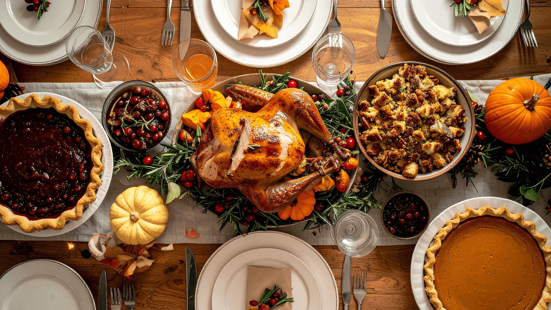 Overhead view of a festive holiday dinner table featuring roasted turkey, pumpkin pie, cranberry pie, stuffing, and seasonal decorations.