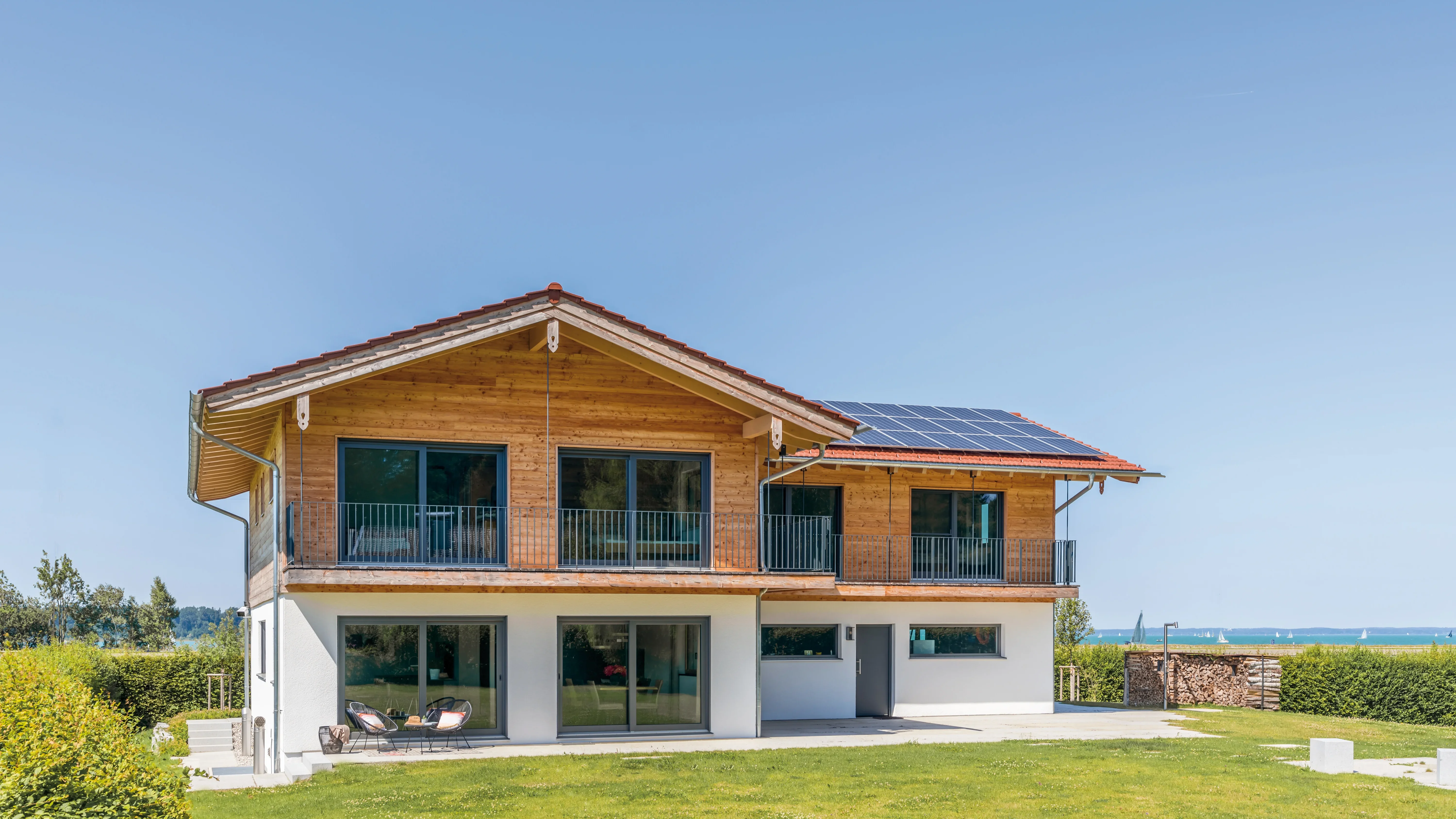 Modernes Holzhaus mit Solaranlage auf dem Dach, großzügiger Glasfront und Blick auf einen See mit Segelbooten.