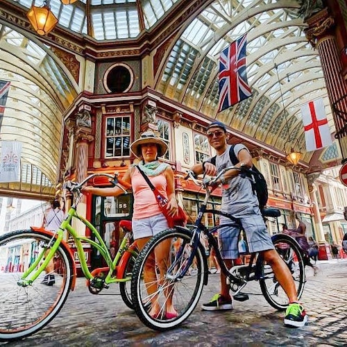 Two people with bicycles stand in a historic, ornate indoor market, adorned with British and English flags hanging from the arched ceiling.