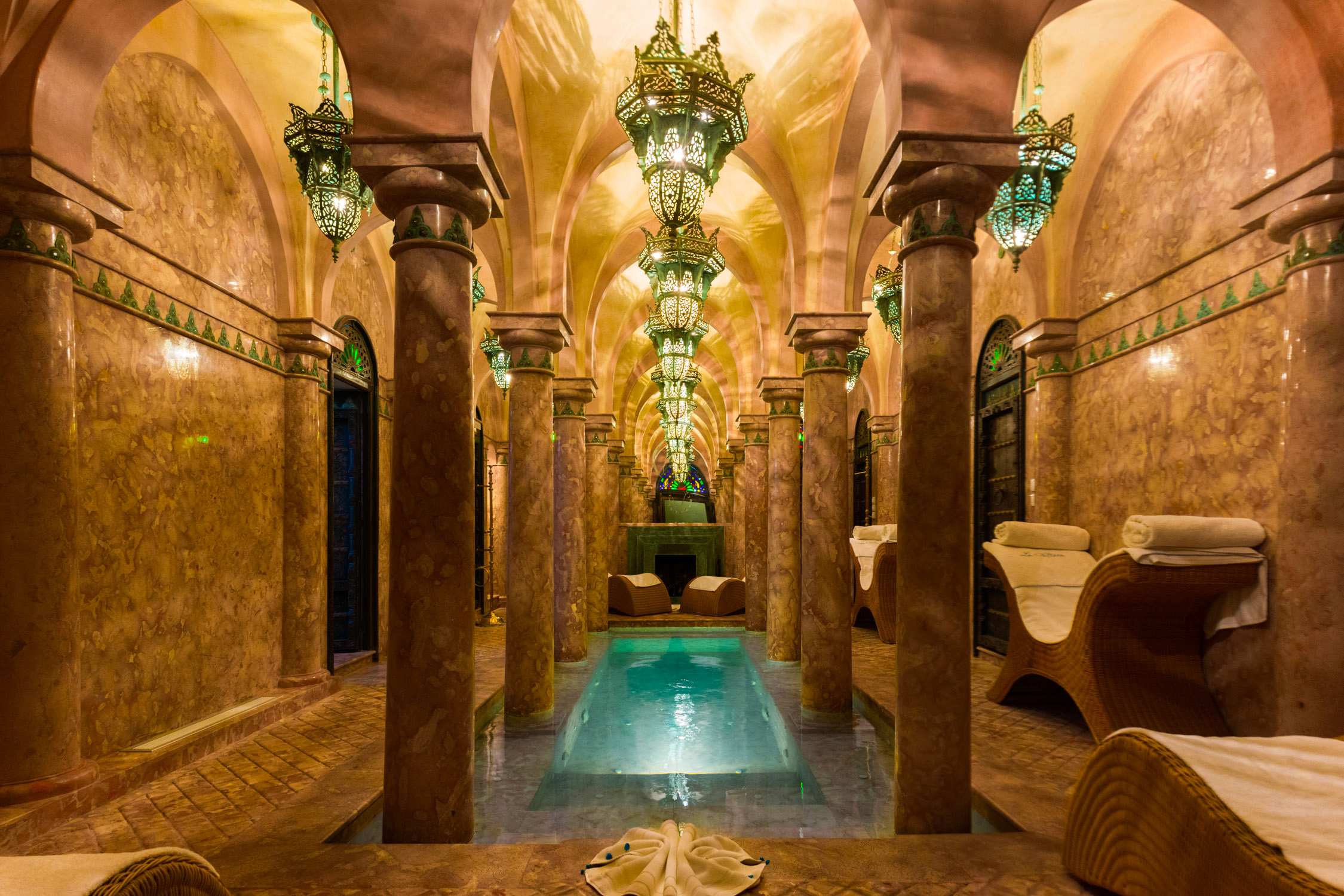 A luxury spa interior with marble columns, mosaic tile walls, and turquoise water in a pool under ornate chandeliers.