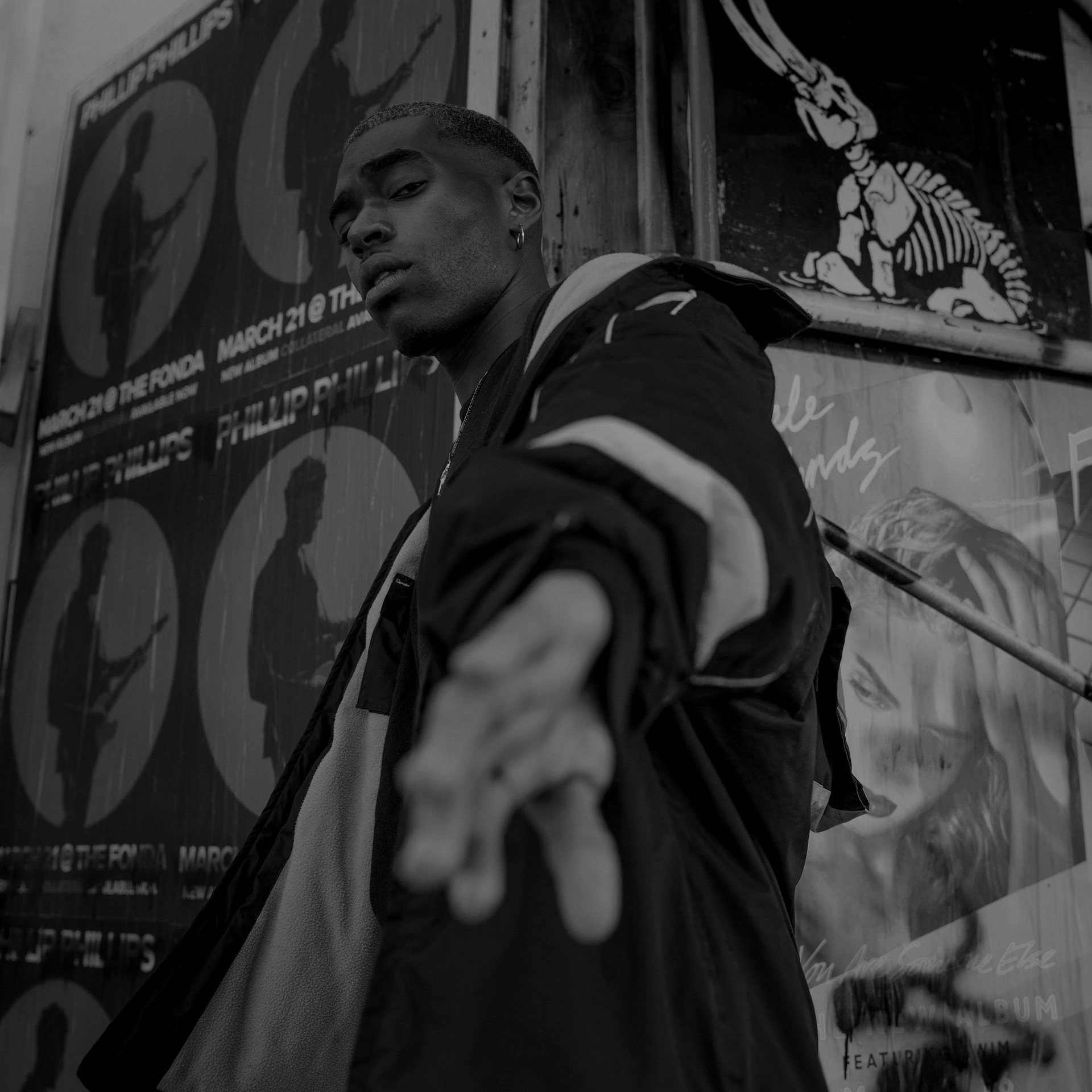 Young man reaching his hand toward the camera, standing against a wall covered in concert posters.