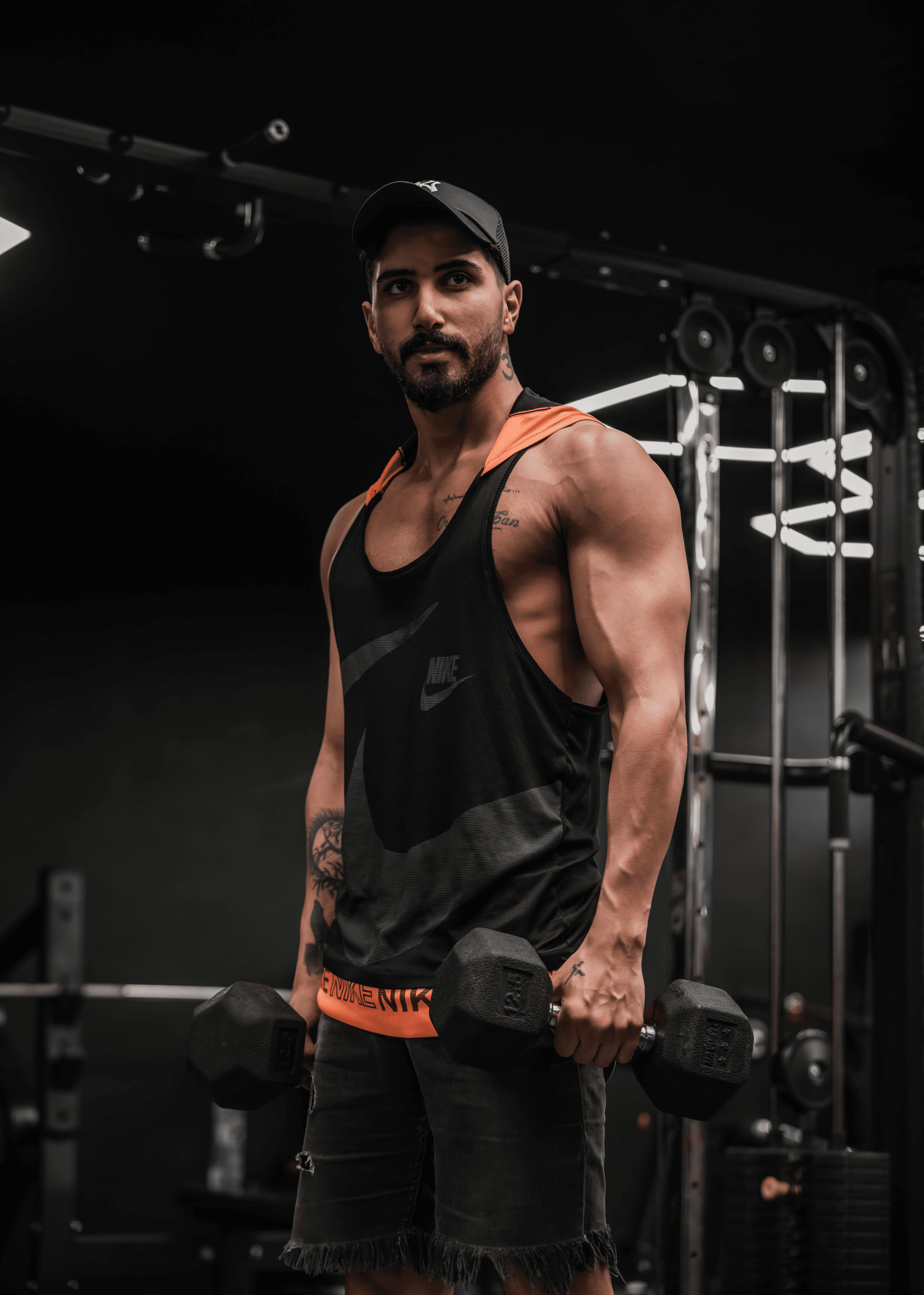 A muscular individual in a gym is lifting a pair of dumbbells, wearing a black tank top and a cap, surrounded by exercise equipment, in a focused bodybuilding workout setting.