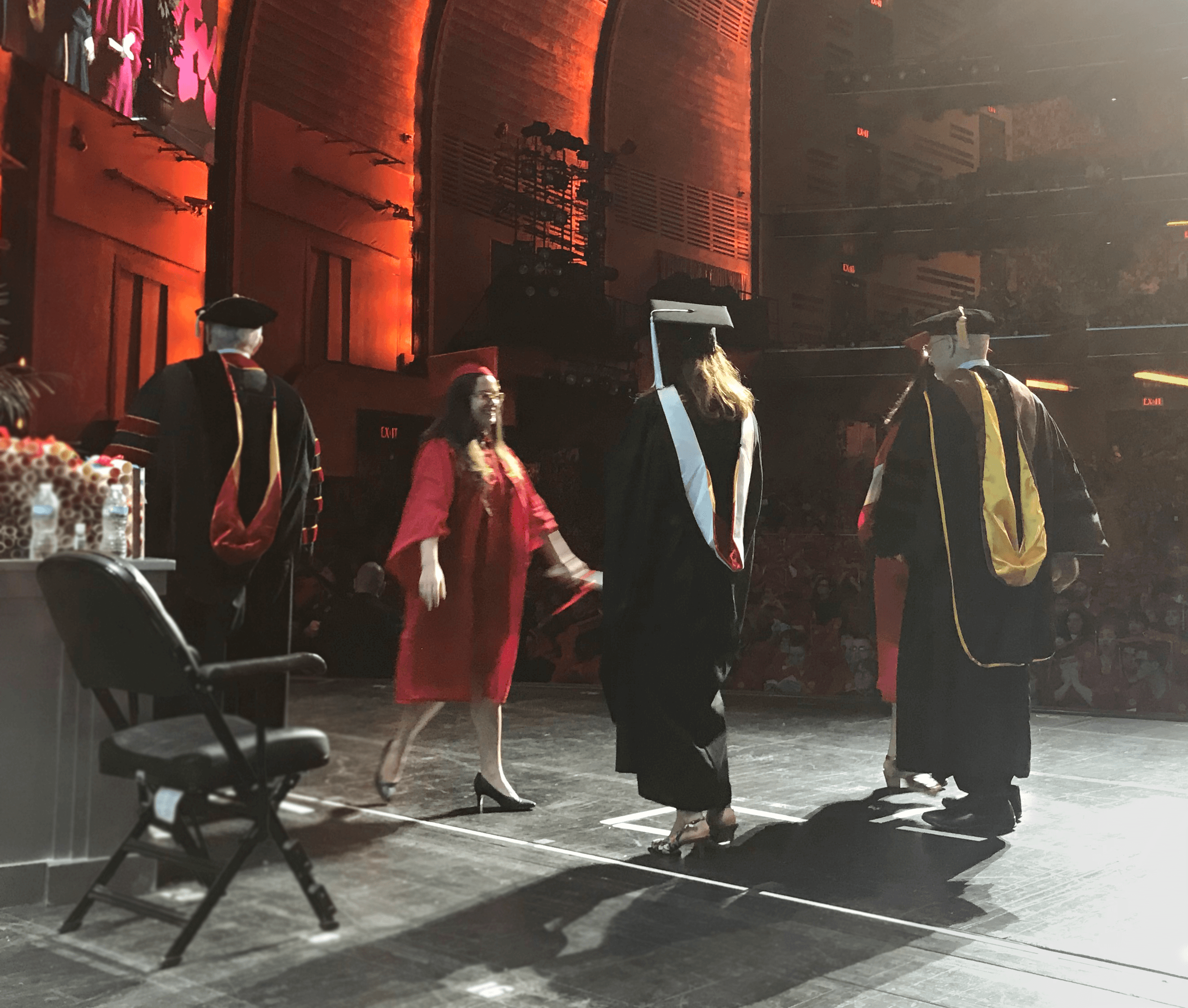 A photo of a group of students and teacher on a stage at the graduation ceremony. They are wearing black or red robes and hats.