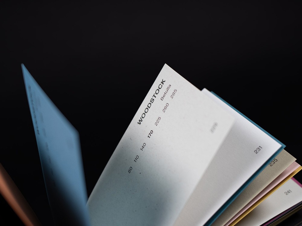 Een sfeervolle detailopname van een opengeslagen waaier van papierstalen. De focus ligt op een witte pagina met de tekst "WOODSTOCK Betulla" en diverse grammages (80, 110, 170, etc.). Op de achtergrond zijn gekleurde randen van andere papierstalen zichtbaar tegen een donkere achtergrond.
