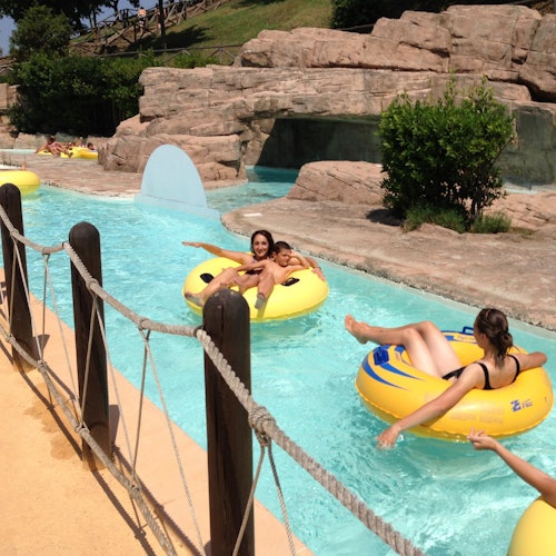 Persone che si rilassano su ciambelle gialle galleggiano lungo un fiume tranquillo con un paesaggio roccioso in una giornata di sole.
