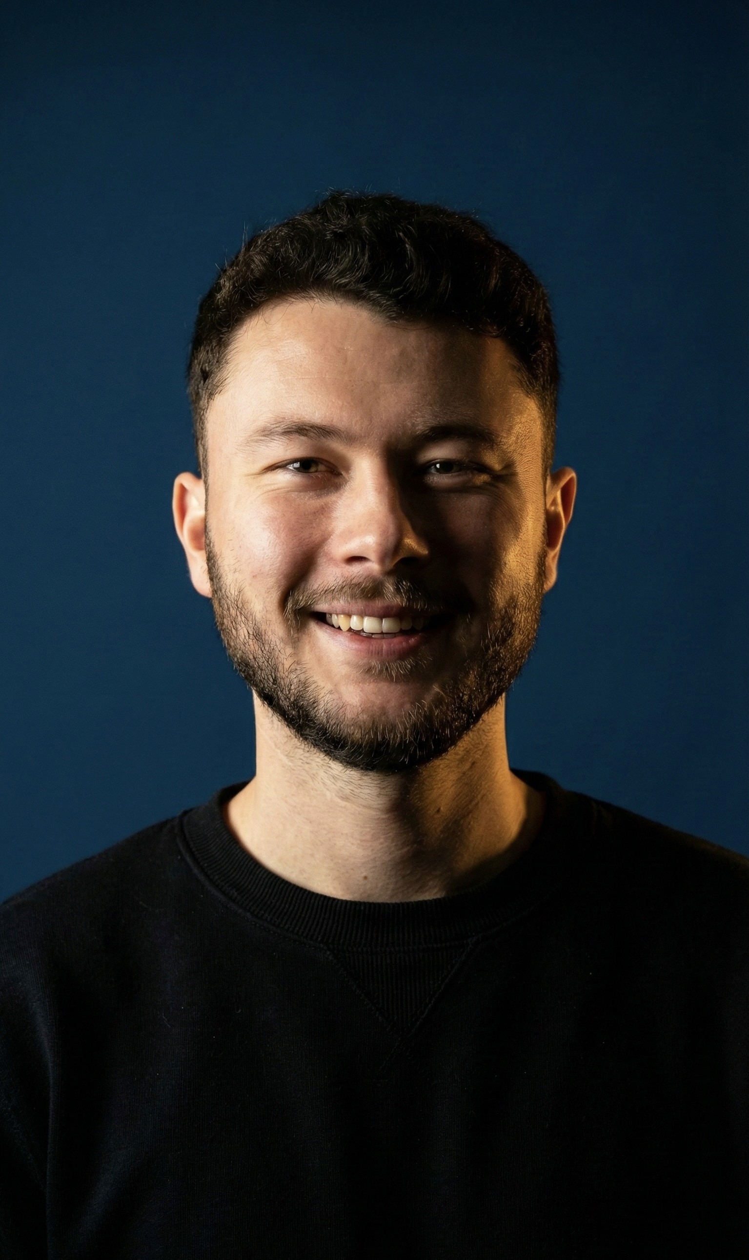 A young woman with long hair smiles while standing against a green backdrop, wearing a casual dark sweater.