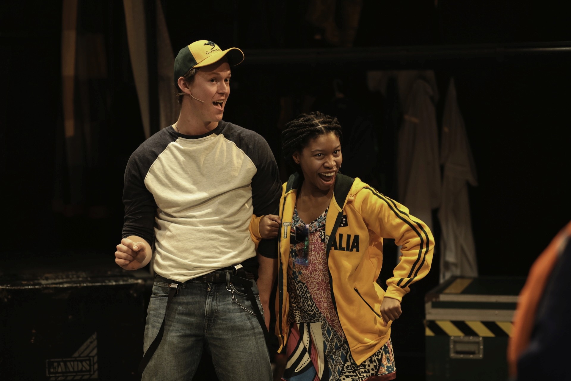 Two performers pose enthusiastically: one in a baseball-style shirt and cap, and the other in an “Australia” jacket, both smiling and mid-gesture in a lively onstage moment.