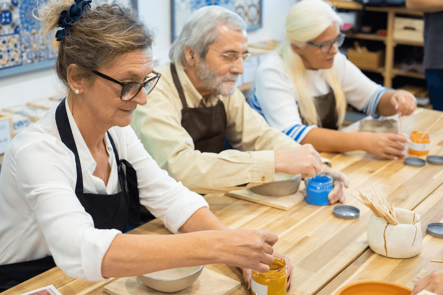 Three older adults in aprons painting pottery side by side at a workshop table.
