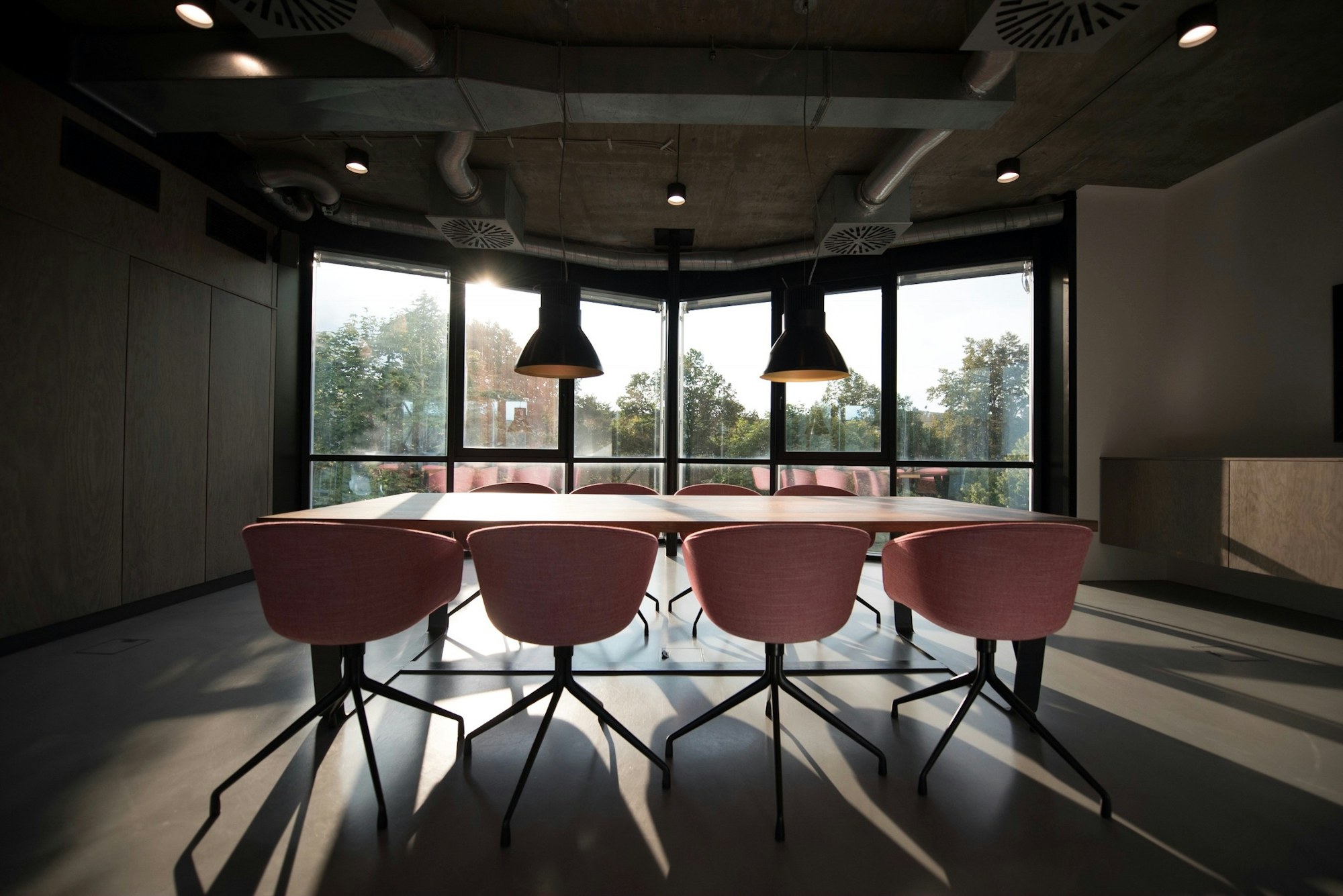 A dark boardroom with pink chairs around a long table and windows behind