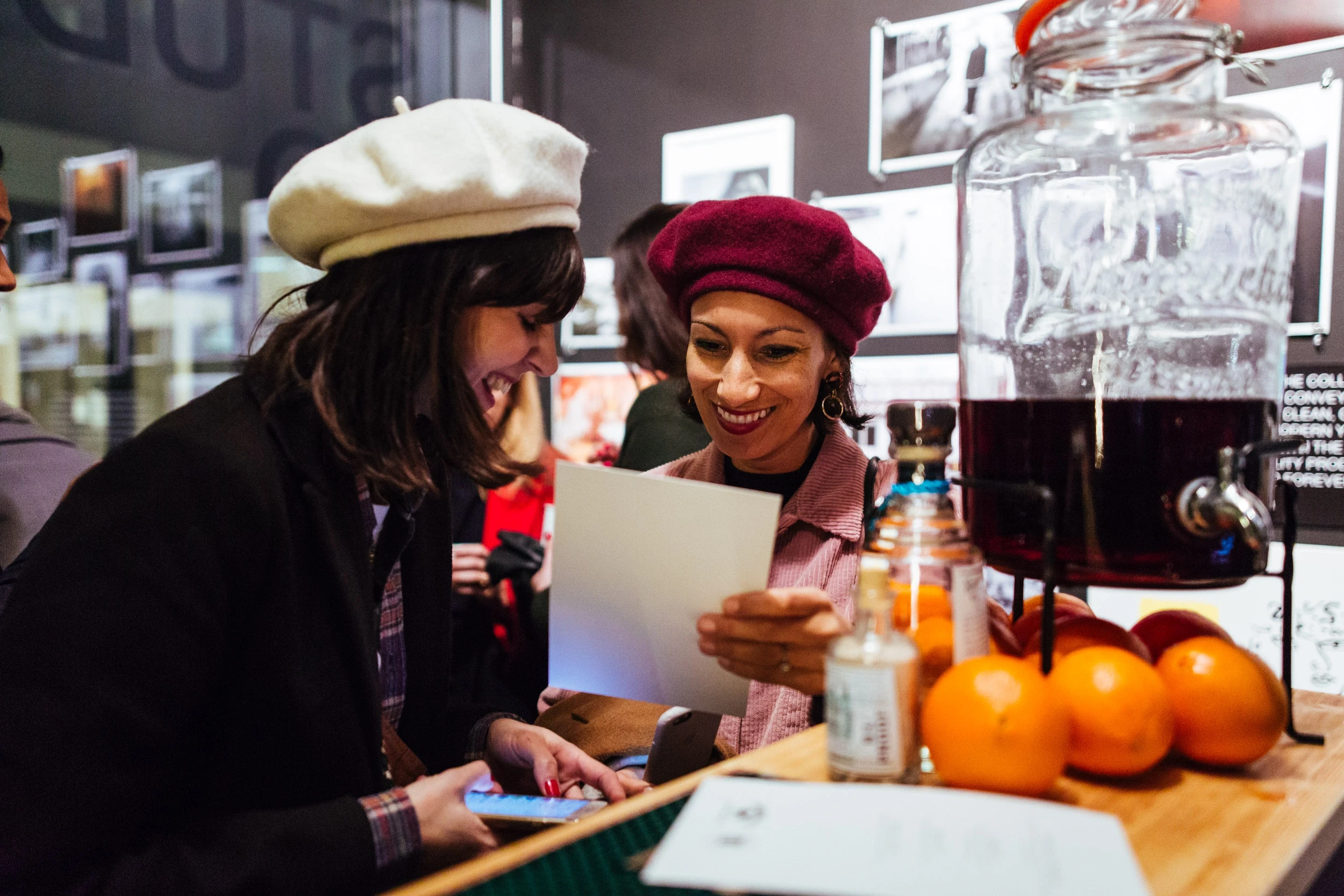 Two smiling guests in berets reading a card at the BRIXTON pop-up bar, next to a glass drink dispenser and fresh oranges.
