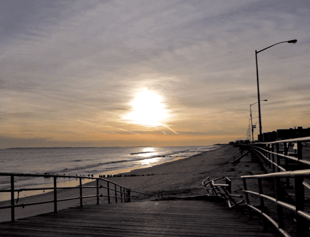 A wooden boardwalk with metal railings extends toward a sandy beach at sunset. The sun is low on the horizon, casting a warm glow over the scene. Streetlights line the boardwalk, and the sky is mostly overcast with light clouds.