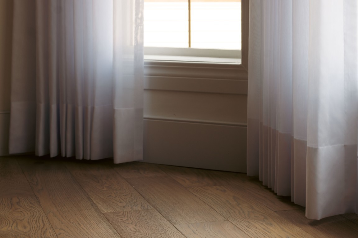 Image of Sheer curtains frame a window with white trim and baseboards in a room with hardwood flooring.