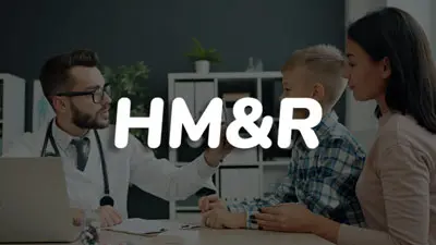 Fotografía de un médico en consulta pediátrica revisando el historial clínico en su computadora, mientras conversa con un niño pequeño y su madre.