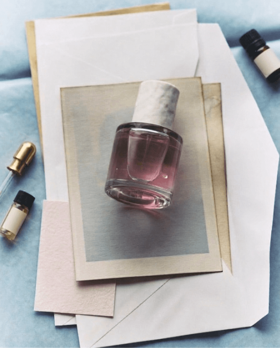 A perfume bottle with a marble cap and blank label rests on layered papers in pastel hues. Nearby are small vials, evoking a calm and elegant mood.