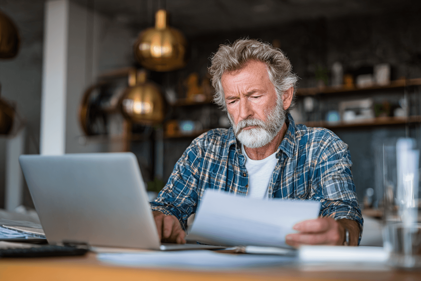 A home care agency owner, male 50s, reviewing financial documents on his laptop at his desk in his office. He looks concerned.
