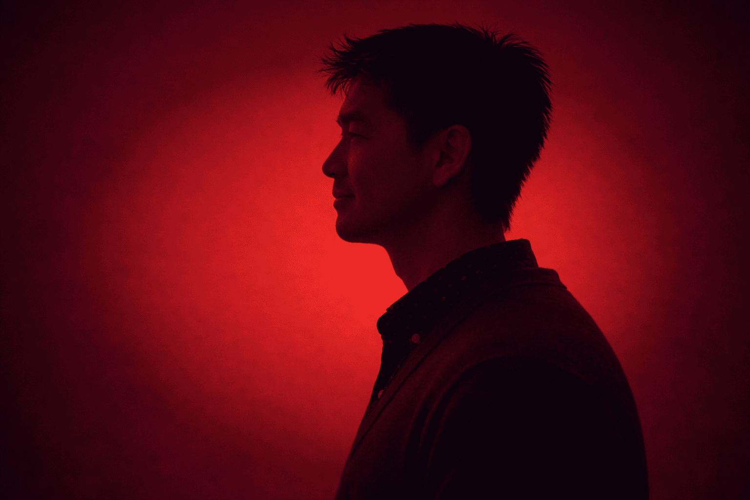 Side-profile portrait of a person facing left, wearing a blazer over a patterned shirt, silhouetted against a vivid red gradient background with soft diagonal shadow bands.