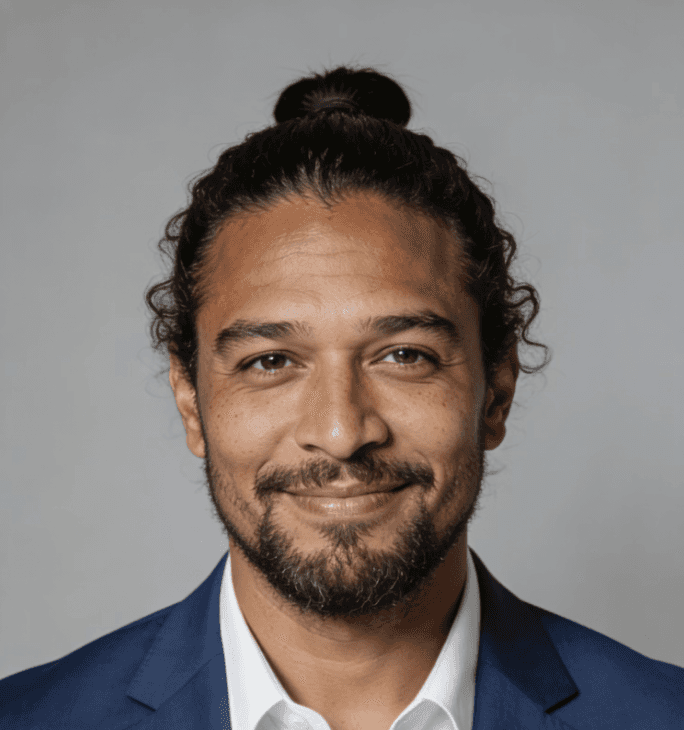 A man with a bun and beard in a blue suit and white shirt smiles warmly against a neutral gray background.
