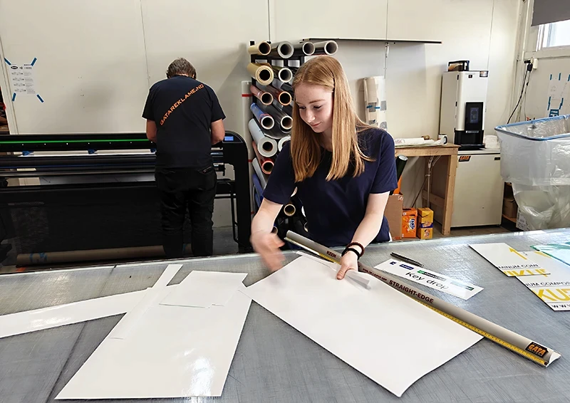 Lærling i profileringsdesign kutter folie presist på arbeidsbord, omgitt av verktøy og fargerike materialer.