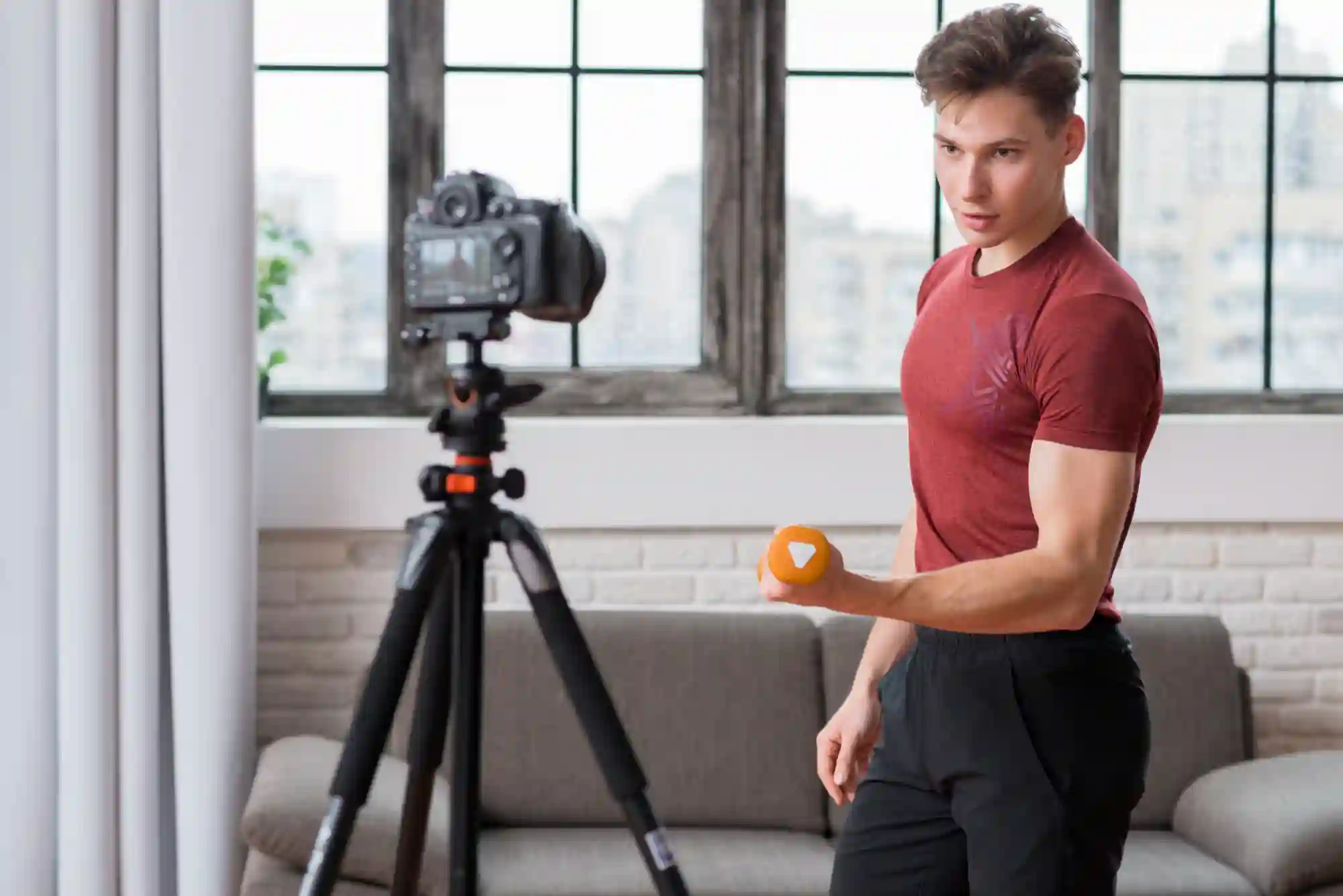 A fit man filming a home workout video with a camera on a tripod while lifting a dumbbell.