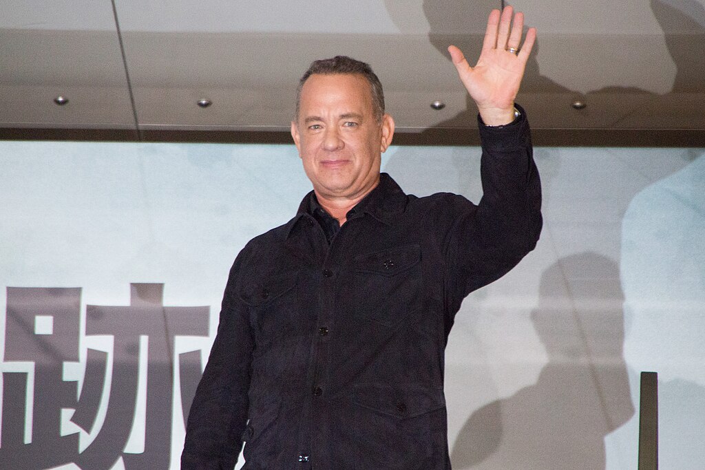 Tom Hanks in a dark jacket waves with his left hand. He stands in front of a geometric patterned background with some partially visible text.