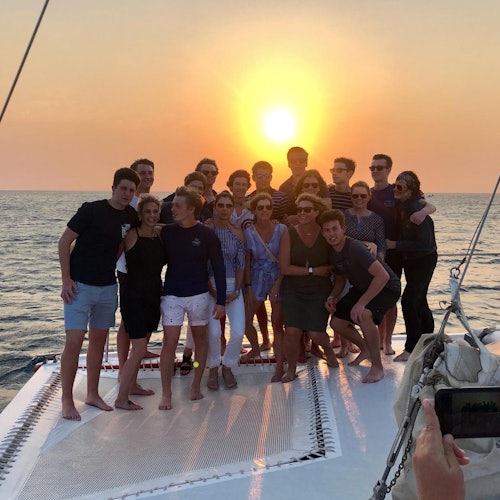 Un groupe de personnes posant sur le pont d'un bateau au coucher du soleil, avec l'océan en arrière-plan et une personne prenant une photo.