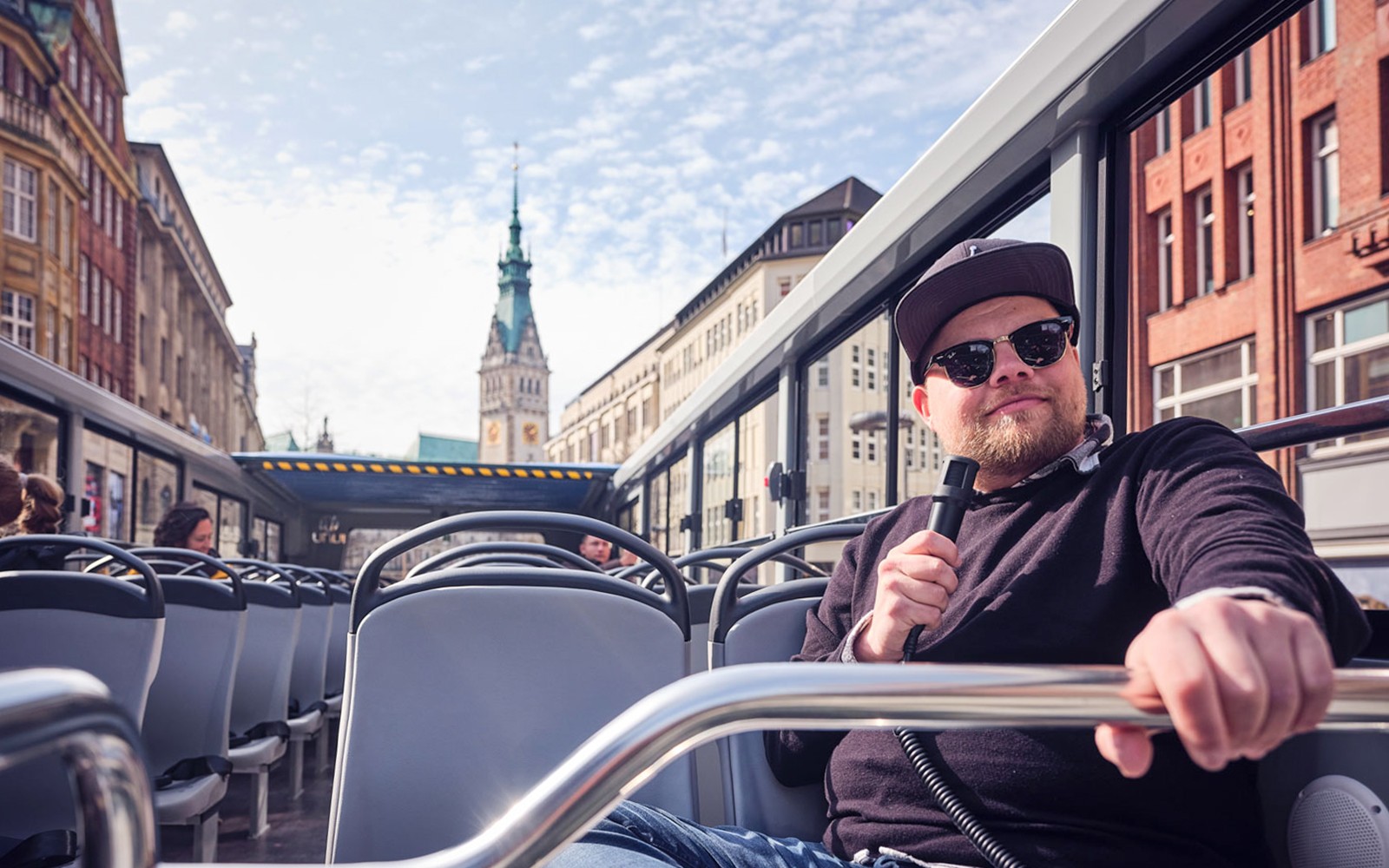 Hop-On Hop-Off bus tour guide in Hamburg with cityscape in background.
