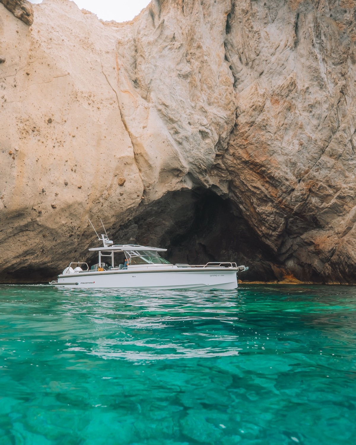 White Axopar 37 yacht anchored in crystal-clear turquoise waters beneath dramatic limestone cliffs and natural rock arch formations in the Greek islands.