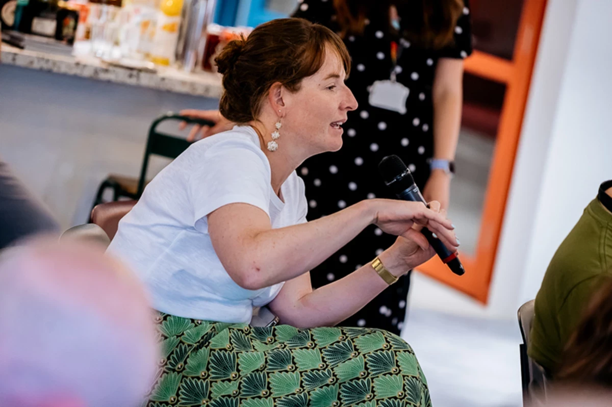 Person seated and holding a microphone during a discussion, wearing a white top and a green patterned skirt, with a countertop and refreshments visible in the background.