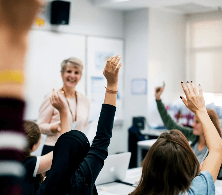 A classroom scene with a teacher standing at the front near a whiteboard, while several students seated at desks raise their hands. Laptops and classroom materials are visible, with daylight coming in through windows.