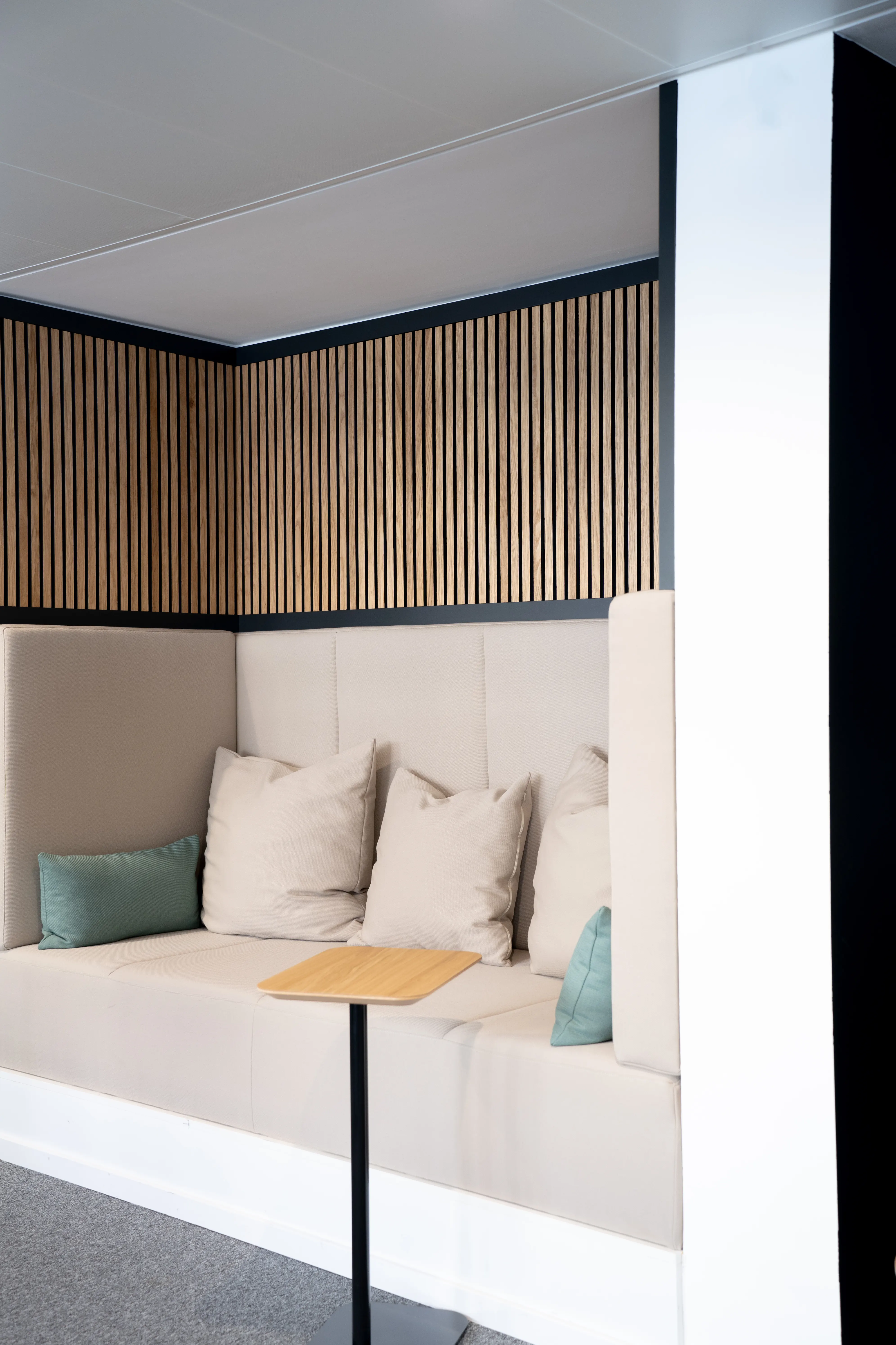 Quiet alcove at AI START-UP MUNICH with a built-in cream upholstered bench, beige and mint cushions, oak slat panelling above and a black standing table with wooden tray.
