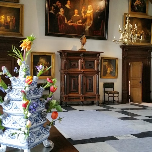 En dekorativ porcelænsblomsteropsats står på et bord i et elegant rum med klassiske malerier, en lysekrone og træmøbler.