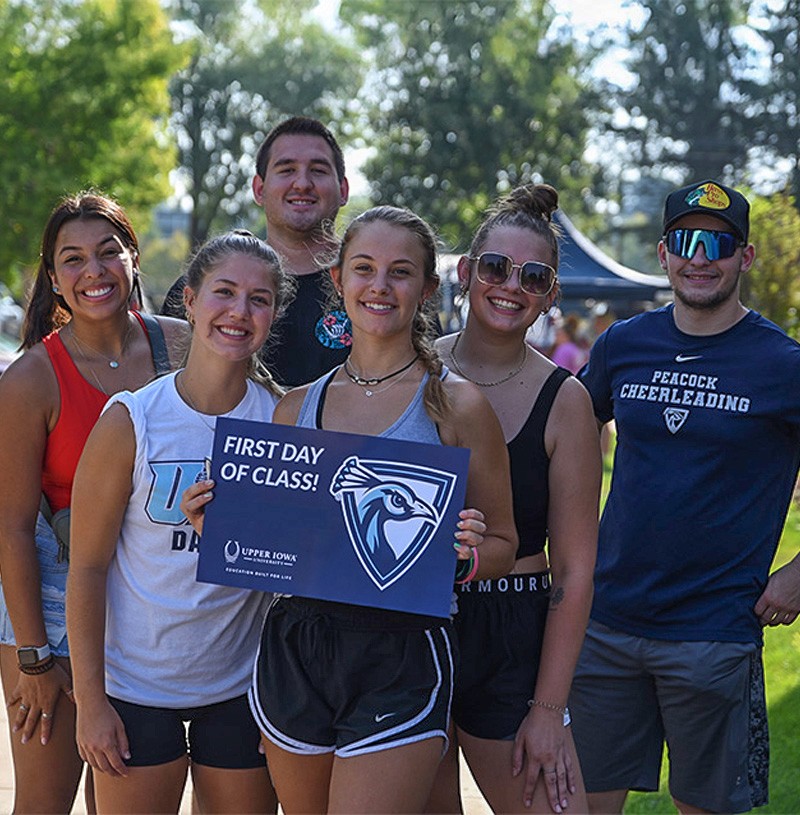 A group of UIU students pose for the camera on a sunny day. One student holds up a "first day of class" sign
