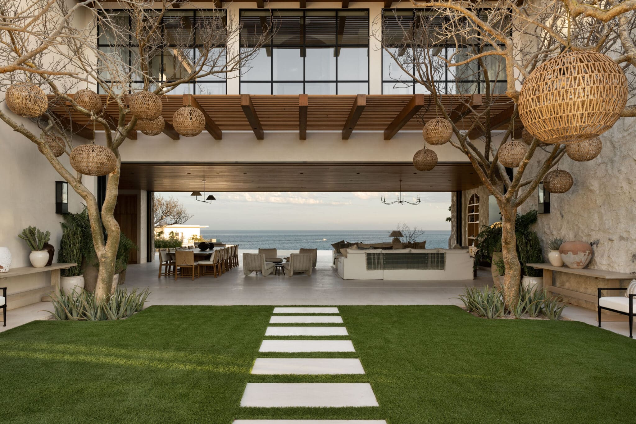 Luxury oceanfront patio with covered outdoor living space, featuring large woven pendant lights hanging from wooden beams. A concrete stepping stone path crosses a manicured lawn, leading to a covered seating and dining area with ocean views. Bare branched trees frame the space, with modern outdoor furniture and built-in features visible in the background.