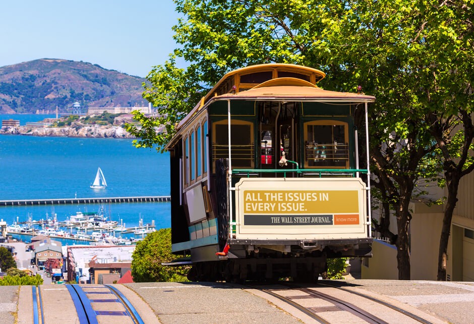 A classic San Francisco cable car travels uphill with a view of the bay, Alcatraz Island, and a sailboat in the background, capturing the core truth and brand authenticity of this iconic city scene.