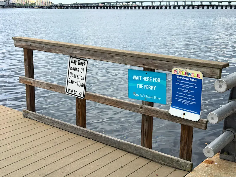The image shows a wooden dock at Bradenton Riverwalk, FL, featuring signs with dock hours, ferry wait instructions, and day dock rules, overlooking the serene waterfront with a distant bridge and cityscape.