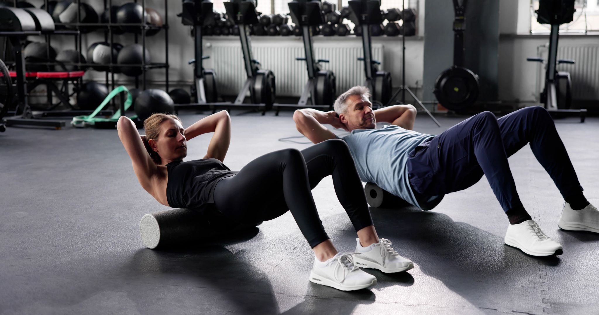 A man and a woman in a gym using foam rollers to roll out their upper backs.