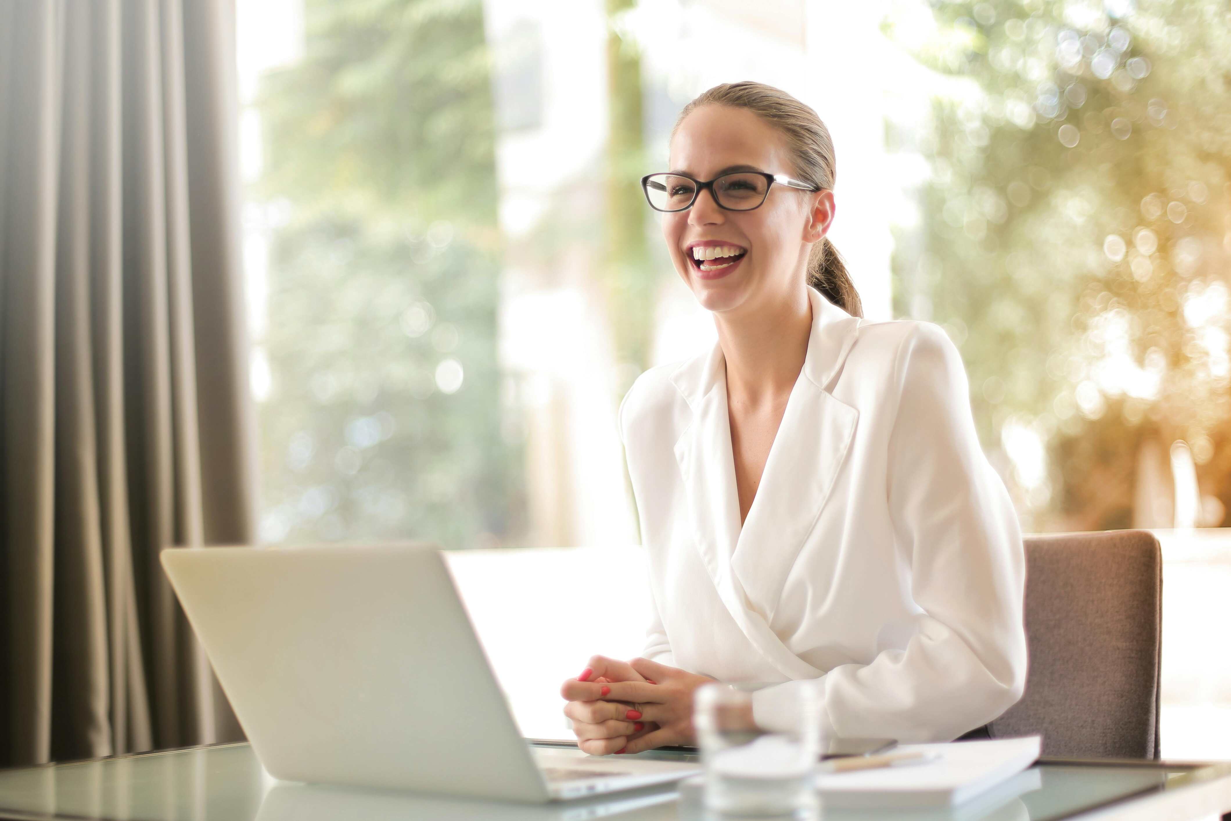 Smiling professional woman in a white blazer sitting at a desk with a laptop, offering a welcoming and expert consultation.