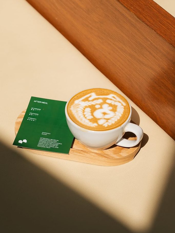 A beautifully crafted latte with intricate latte art, placed on a wooden tray alongside a green menu card, set against a minimalist backdrop.