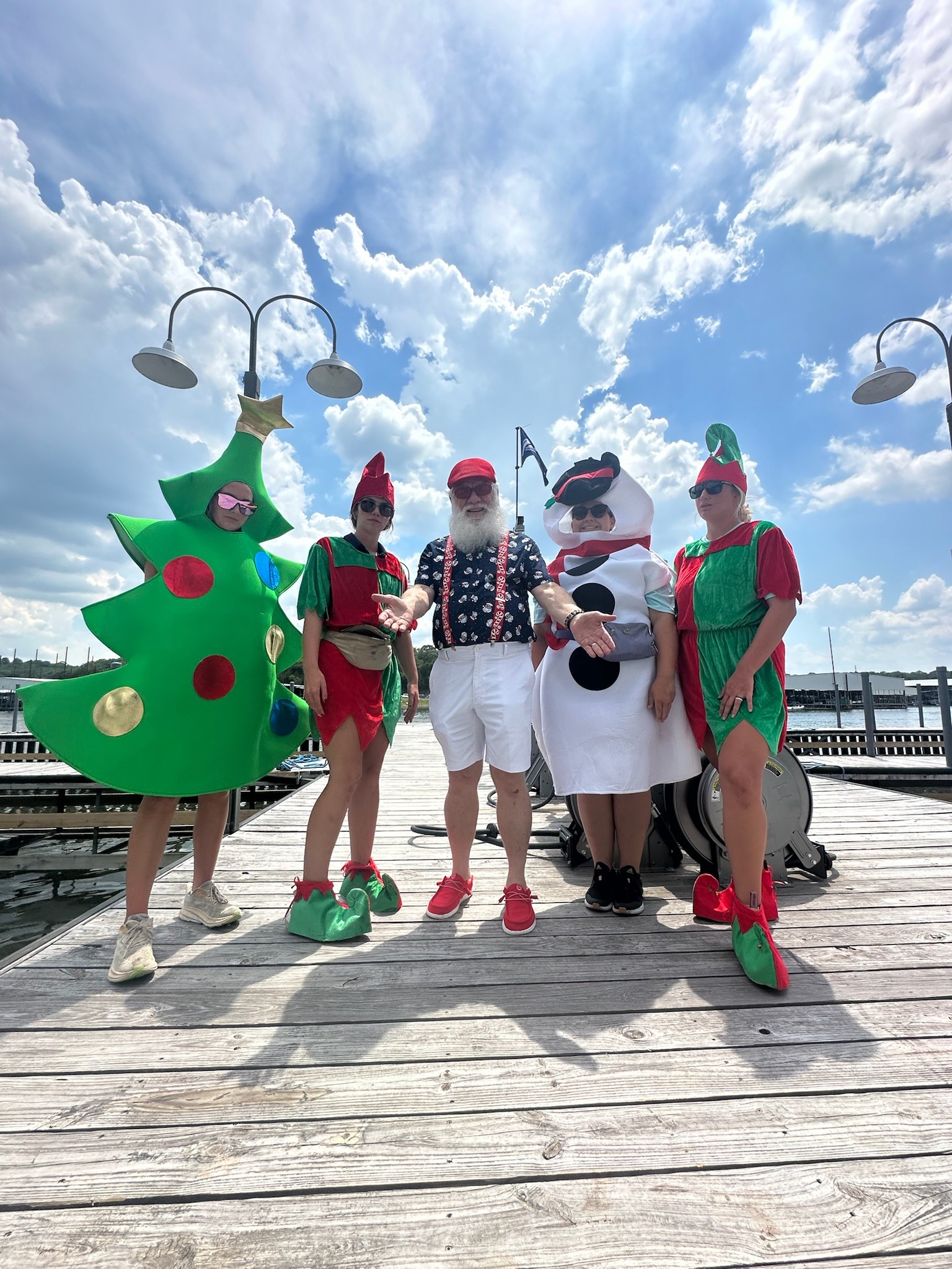A group of people stands on a wooden dock under a partly cloudy sky, dressed in festive costumes including a Christmas tree, elves, and a snowman, with lampposts in the background.