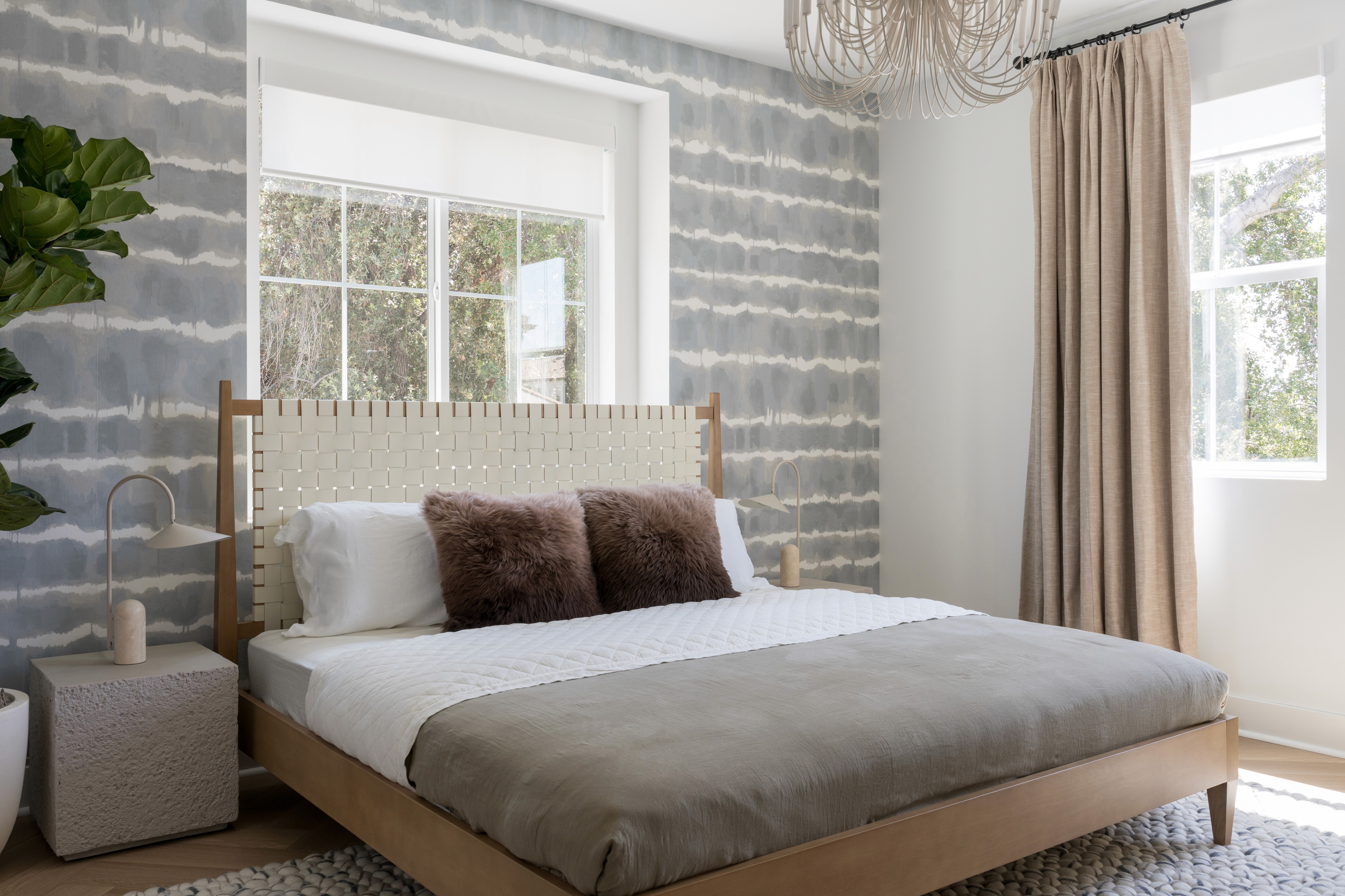 Bedroom with neutral wood bed frame, white, leather woven headboard, square side table with arched table lamp, sage green bedding with brown fur accent pillows, cream curtains, grey and white watercolor accent wall, woven rug, and white, metal chandelier by Jubilee Interiors in Arcadia, California