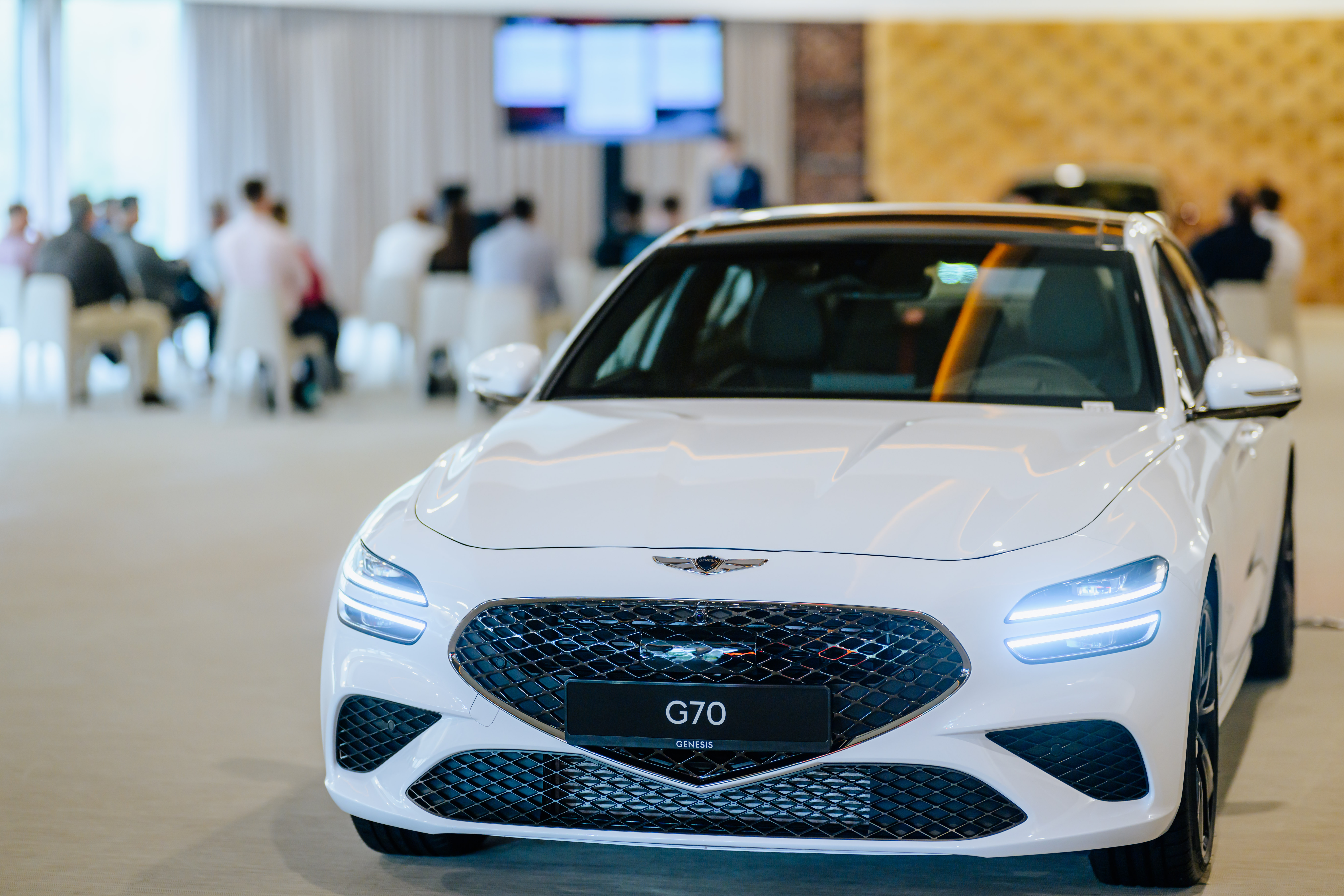 Luxuslimousine Genesis G70 in weißer Ausführung im Vordergrund eines exklusiven Automobil-Events mit Präsentation im Hintergrund.