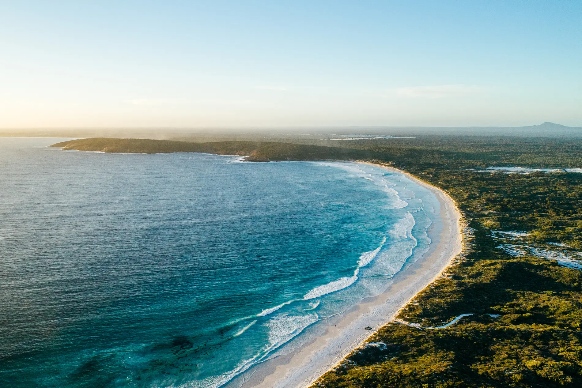 Aerial at dusk of a long beach curving away beneath low coastal heath