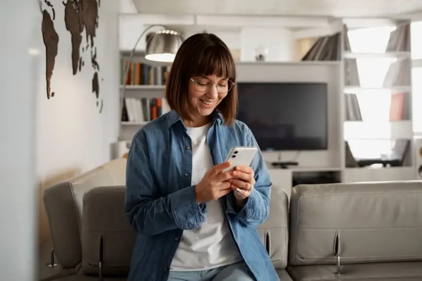 A woman sits comfortably in a modern living room, smiling as she looks at her phone. The image represents a relaxed homeowner exploring refinancing options with Chris Lewis Home Loans, showing how easy and accessible the process can be.