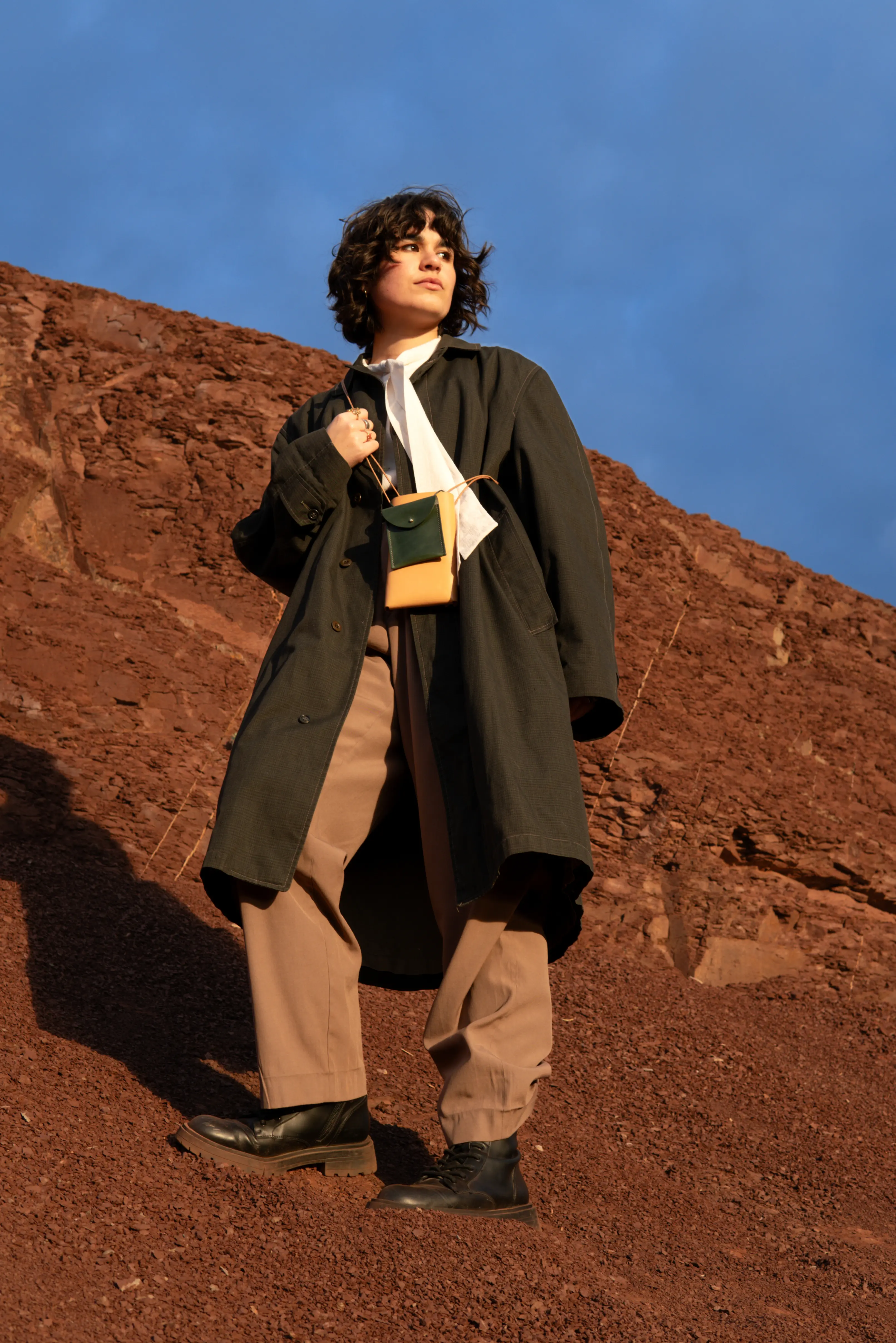 Full-length portrait of Anaïs Brancheriau wearing a dark coat, beige trousers and black boots, standing on a red rocky slope with a two-tone crossbody pouch.